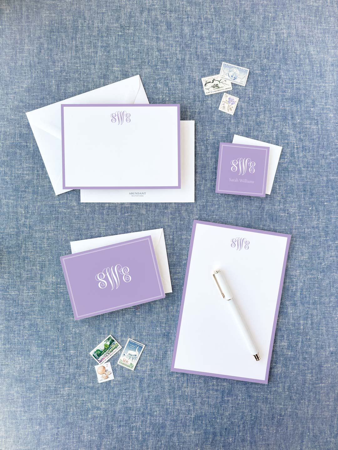Personalized lavender bordered stationery set featuring elegant monogrammed cards, notepad, envelopes, and a white pen on a textured blue background.