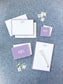 Personalized lavender bordered stationery set featuring elegant monogrammed cards, notepad, envelopes, and a white pen on a textured blue background.