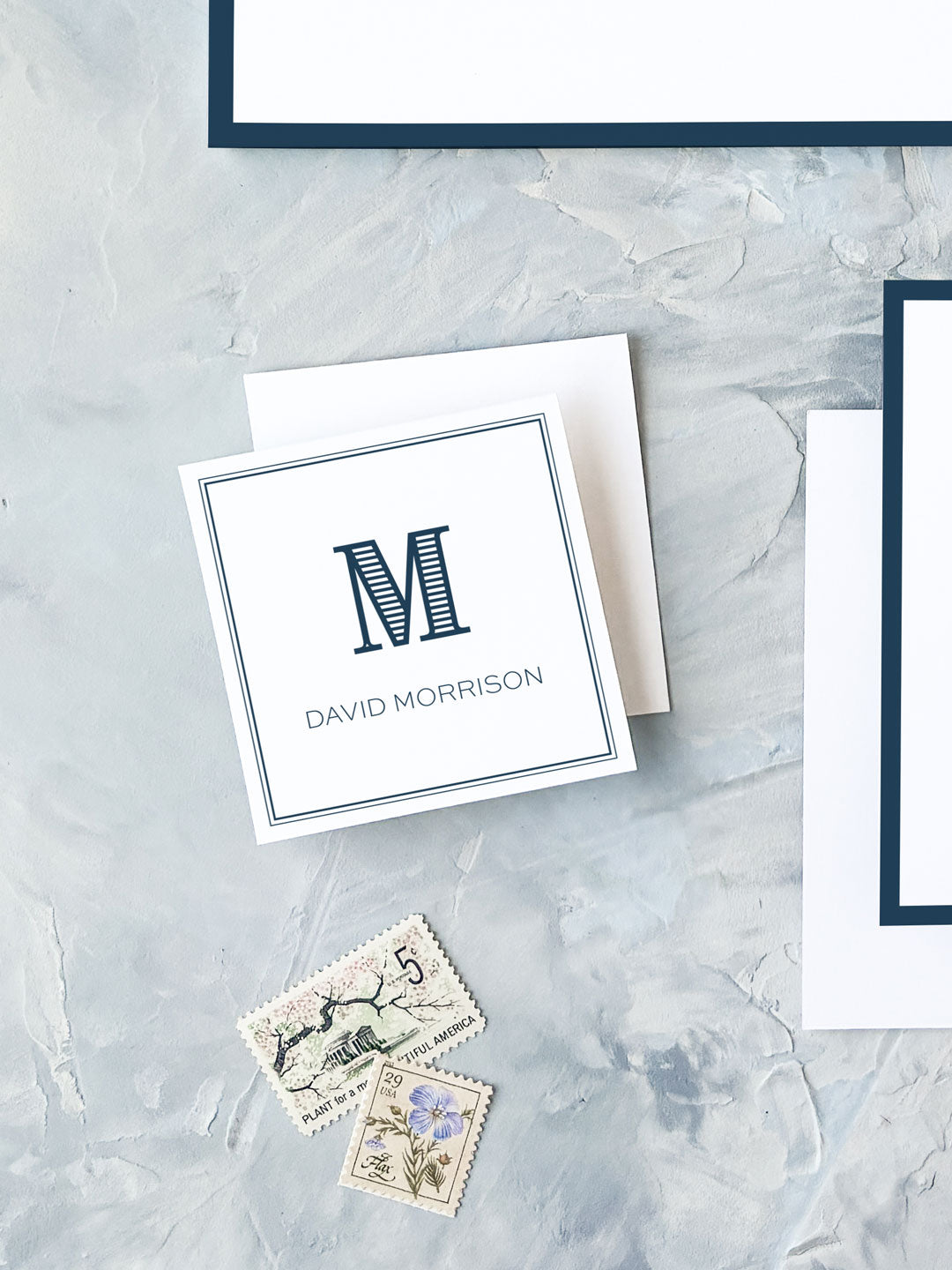Square personalized calling card with a navy monogram letter and name on white textured background, accompanied by vintage stamps.