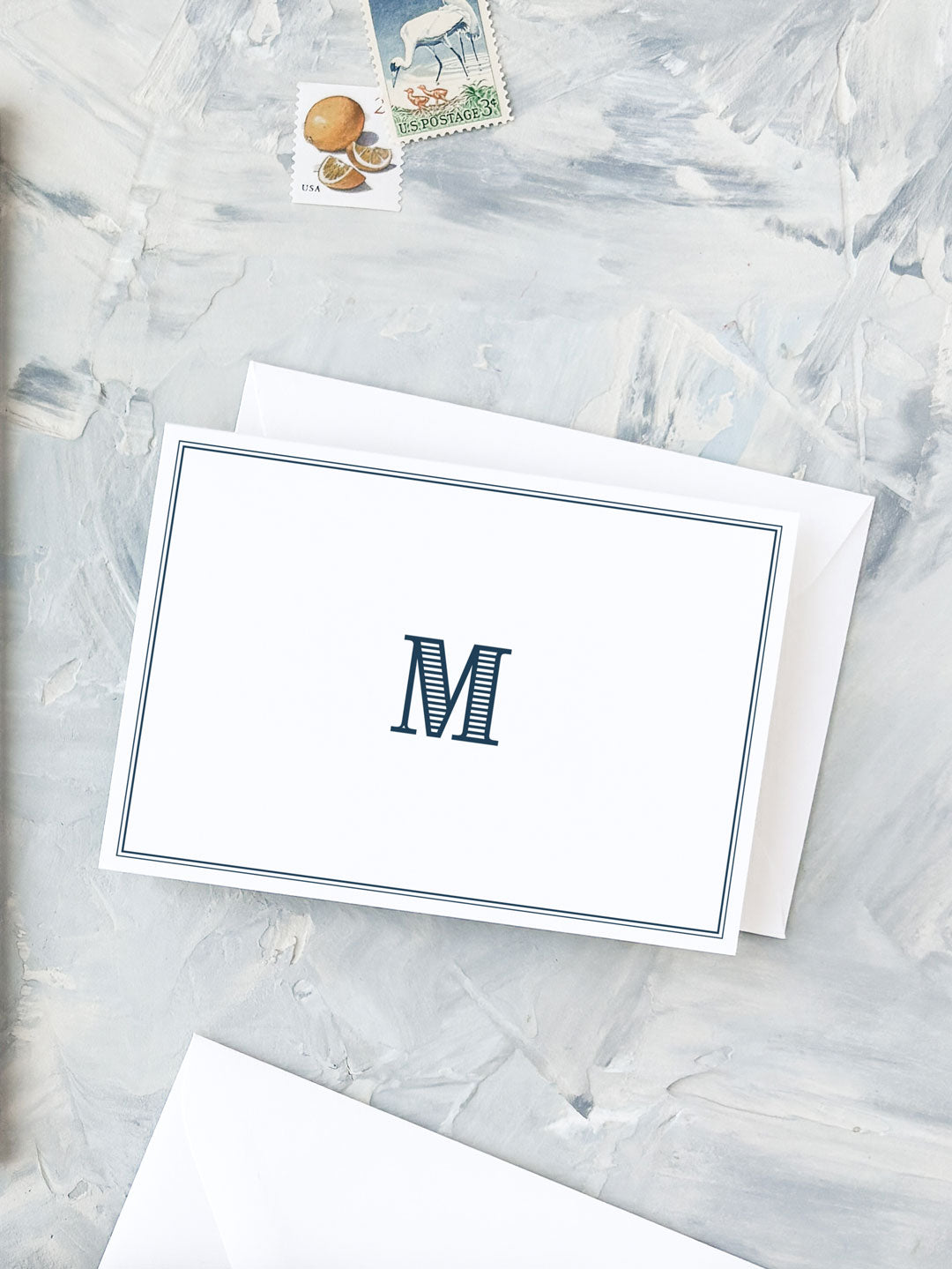 Folded notecard with a navy monogrammed letter "M" framed by a simple double-line border on textured light background with vintage stamps.