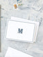 Folded notecard with a navy monogrammed letter "M" framed by a simple double-line border on textured light background with vintage stamps.
