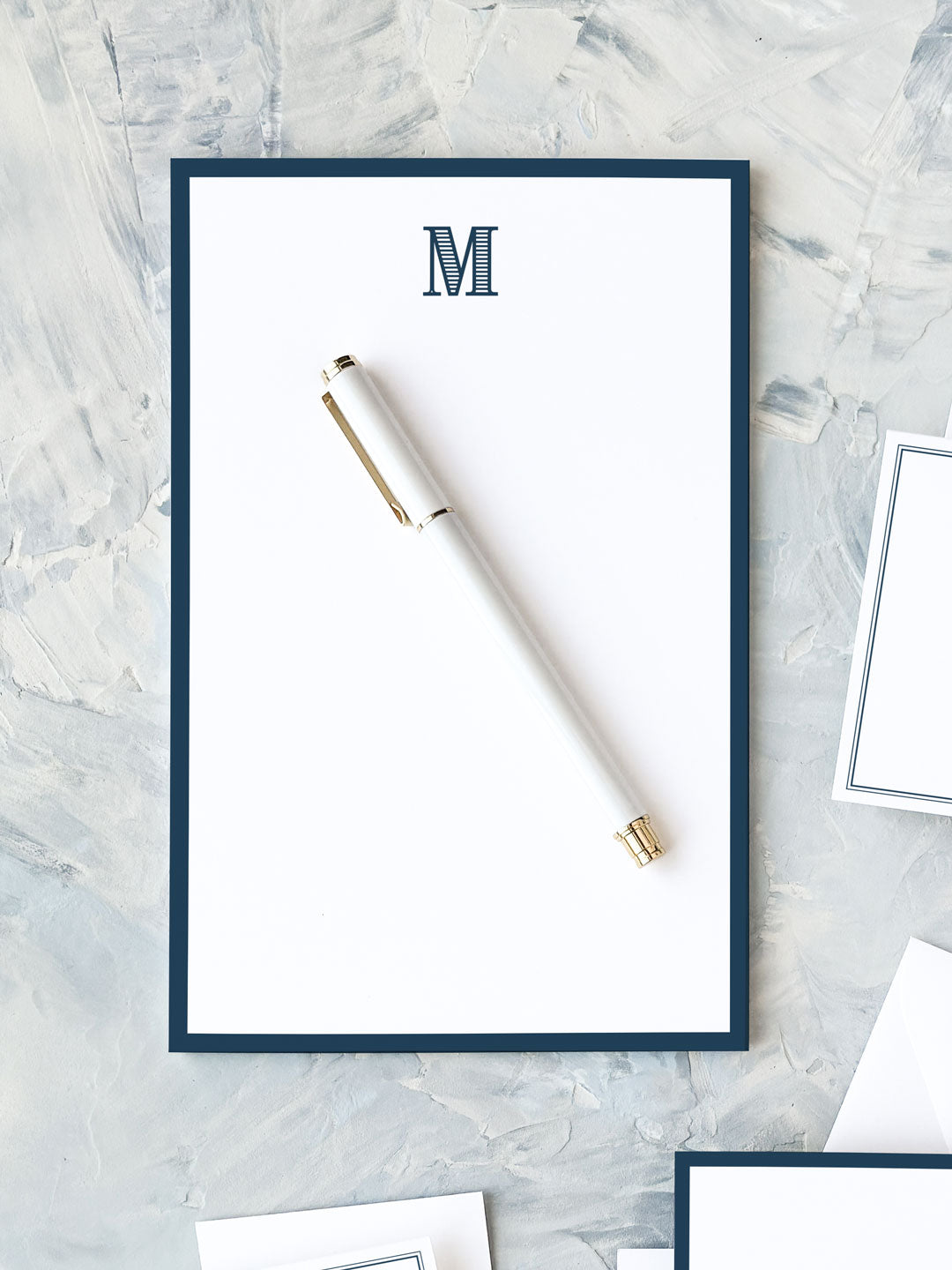 Classic navy bordered notepad with an elegant monogram letter M at the top, displayed with a white and gold pen on a textured gray surface.