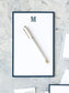 Classic navy bordered notepad with an elegant monogram letter M at the top, displayed with a white and gold pen on a textured gray surface.