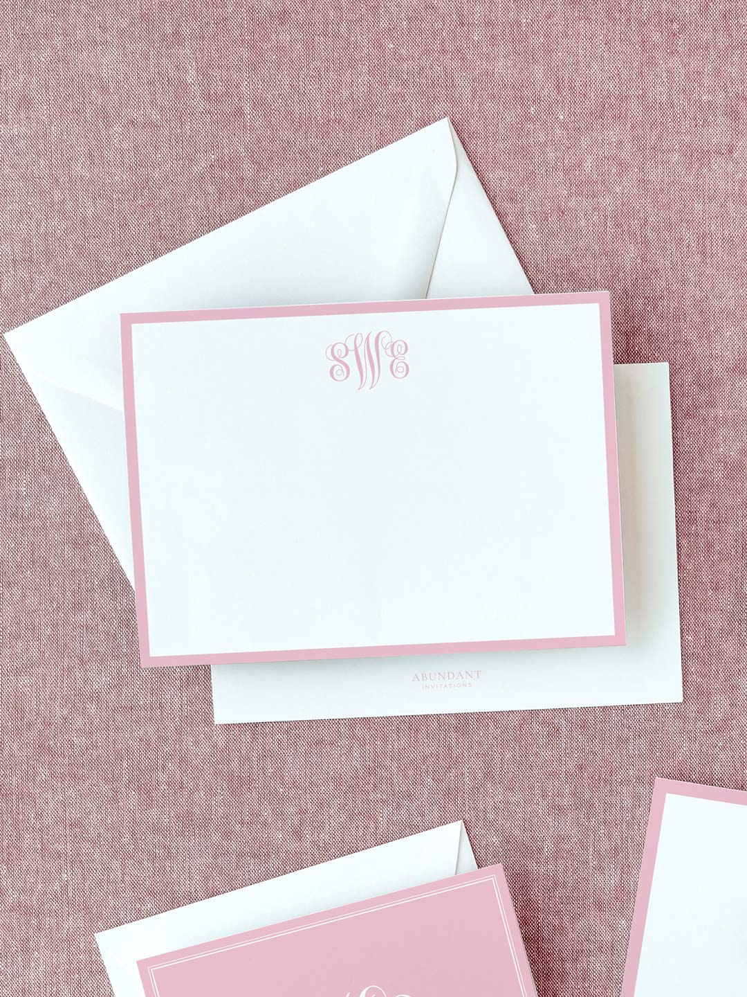 Elegant flat notecard with a soft blush pink border and a decorative monogram at the top, resting on a textured fabric background with white envelopes.
