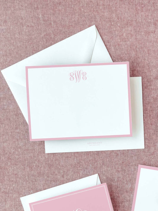 Elegant flat notecard with a soft blush pink border and a decorative monogram at the top, resting on a textured fabric background with white envelopes.