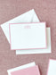 Elegant flat notecard with a soft blush pink border and a decorative monogram at the top, resting on a textured fabric background with white envelopes.