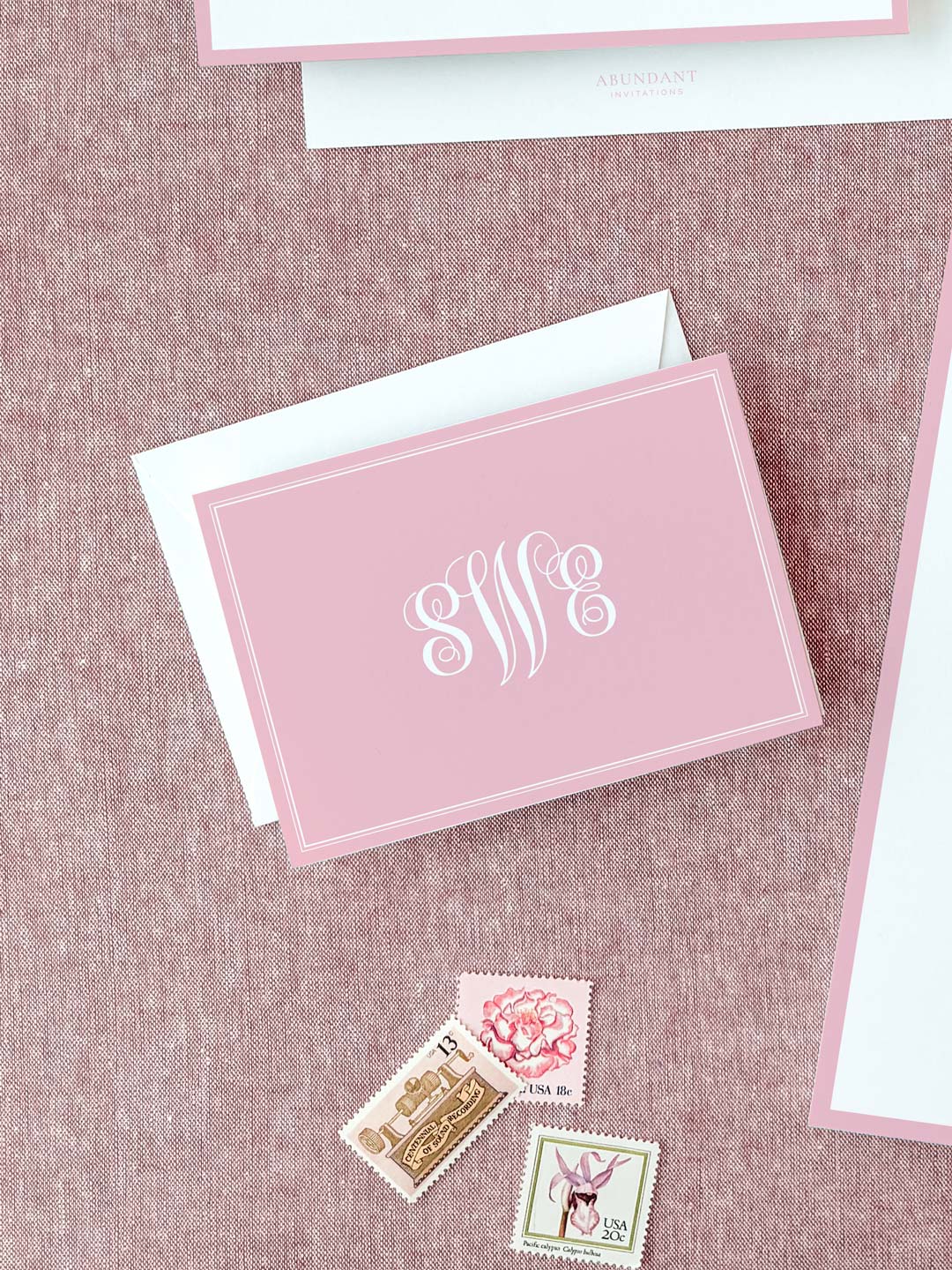Folded blush pink notecard with elegant white monogram lettering resting on textured fabric, accompanied by vintage postage stamps and envelope.