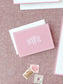Folded blush pink notecard with elegant white monogram lettering resting on textured fabric, accompanied by vintage postage stamps and envelope.