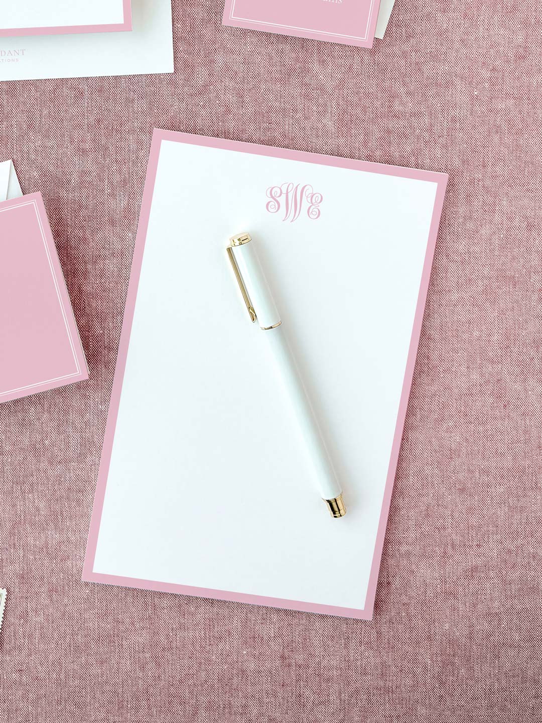 White notepad with soft blush pink border and elegant monogram lettering placed on textured fabric surface with a white and gold pen.