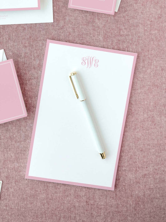 White notepad with soft blush pink border and elegant monogram lettering placed on textured fabric surface with a white and gold pen.