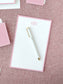 White notepad with soft blush pink border and elegant monogram lettering placed on textured fabric surface with a white and gold pen.