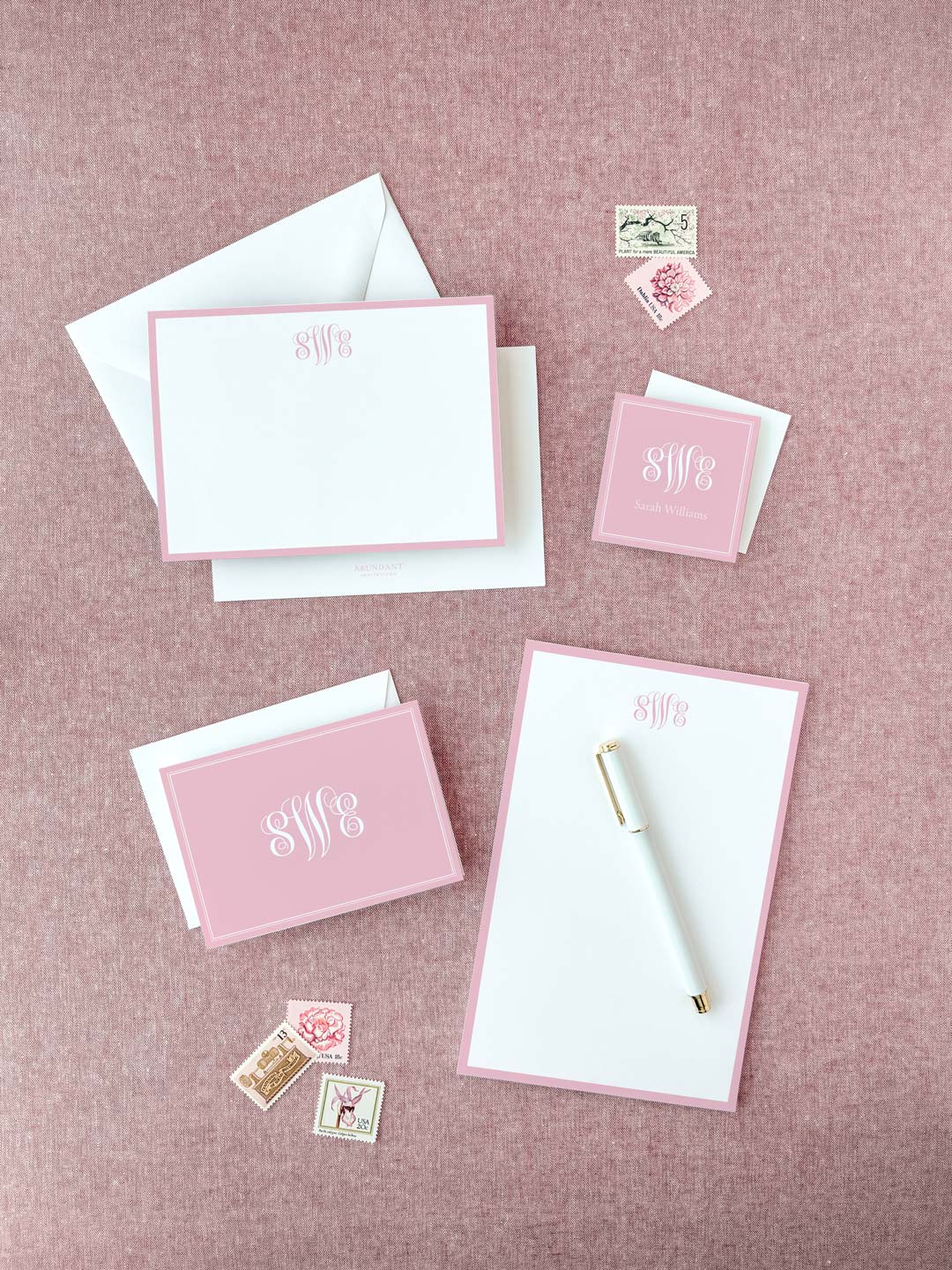 Set of elegant blush pink and white personalized stationery including notepads, envelopes, and coordinating vintage-style postage stamps on textured pink background.