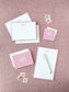 Set of elegant blush pink and white personalized stationery including notepads, envelopes, and coordinating vintage-style postage stamps on textured pink background.