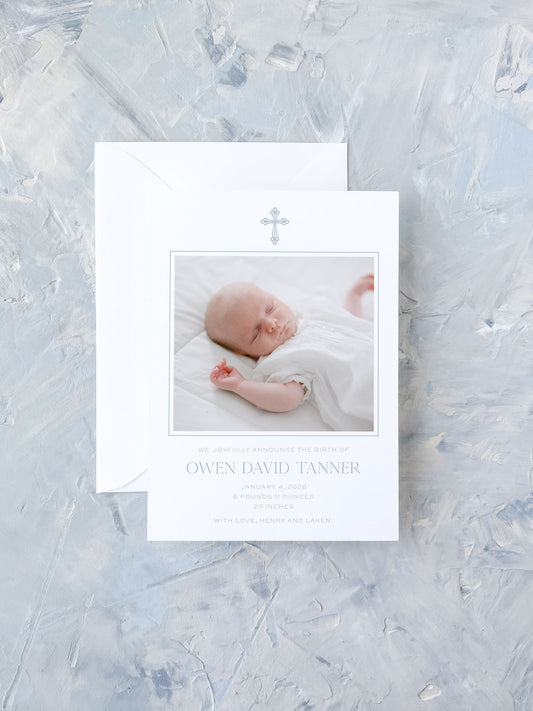 A classic blue-accented birth announcement card featuring a sleeping baby photo and a delicate cross design on white personalized stationery.