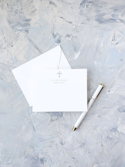 White flat notecard with a subtle blue cross and delicate text, paired with a matching envelope and an elegant white and gold pen on a textured light gray background.