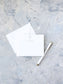 White flat notecard with a subtle blue cross and delicate text, paired with a matching envelope and an elegant white and gold pen on a textured light gray background.