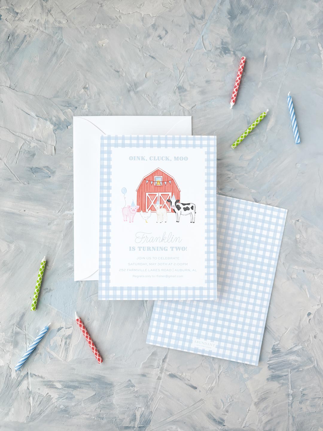 Blue checkered birthday invitation with farm animals and red barn, surrounded by colorful candles on gray surface