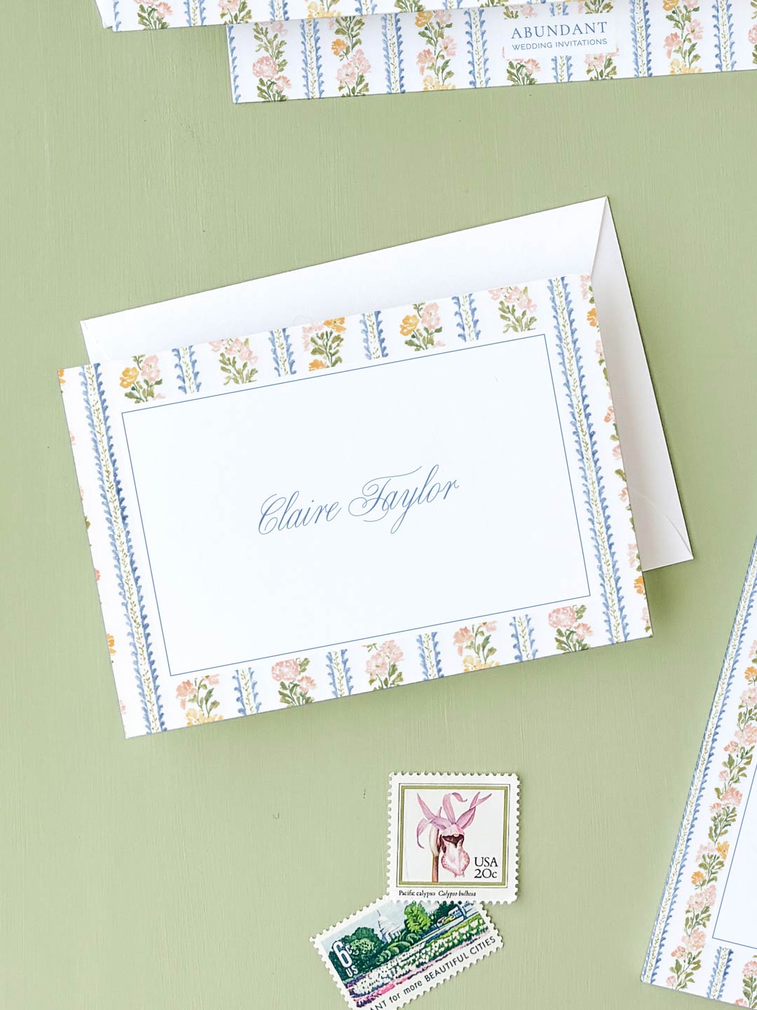 Folded notecard with floral and stripe border design resting on green surface with vintage postage stamps nearby