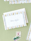 Folded notecard with floral and stripe border design resting on green surface with vintage postage stamps nearby