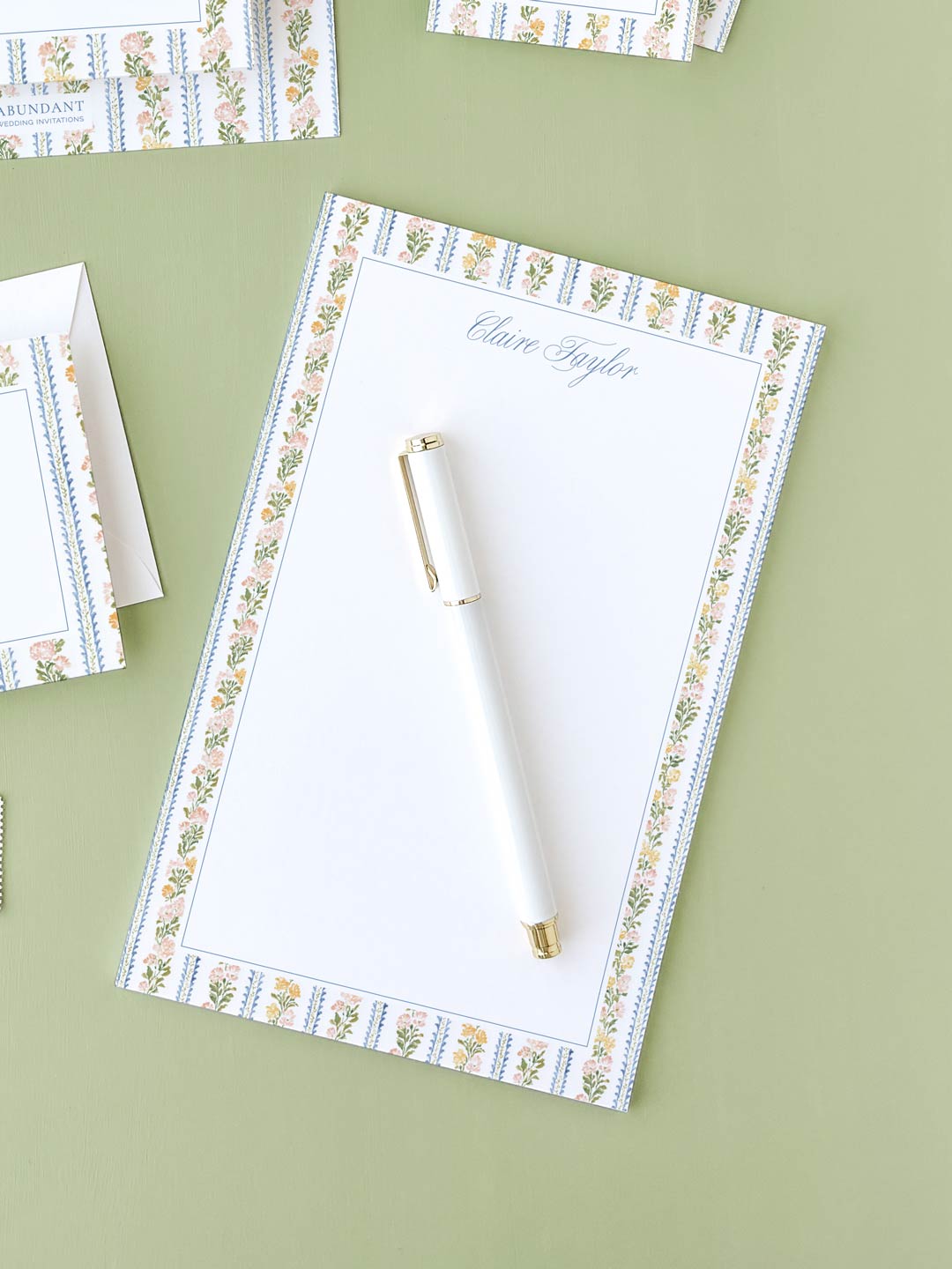 Notepad with botanical stripe border and a white pen placed on a green surface.
