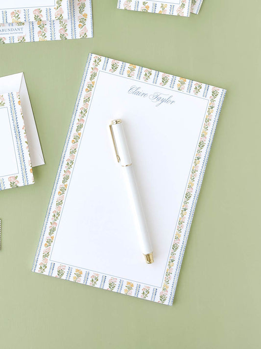 Notepad with botanical stripe border and a white pen placed on a green surface.