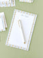 Notepad with botanical stripe border and a white pen placed on a green surface.