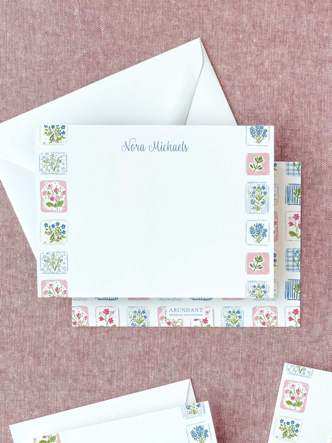 Personalized flat notecard with delicate floral tile border and matching envelope on a textured surface.