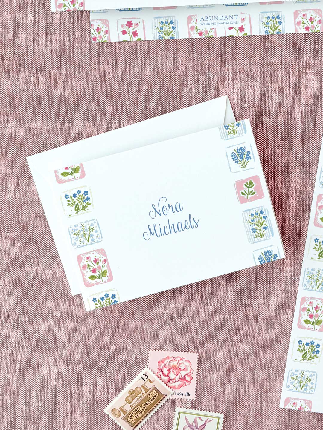 Folded notecard with floral tile border and handwritten name on textured pink background.