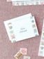 Folded notecard with floral tile border and handwritten name on textured pink background.