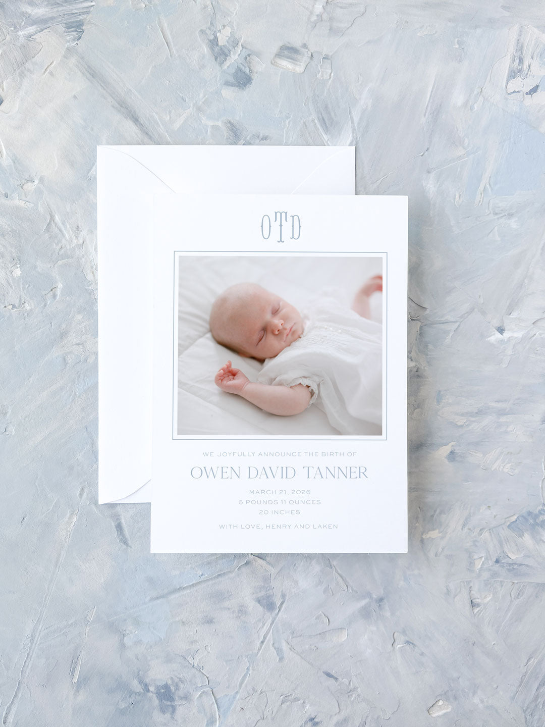 Elegant blue and white birth announcement card featuring a sleeping newborn photo with a monogram and personalized details on textured background.