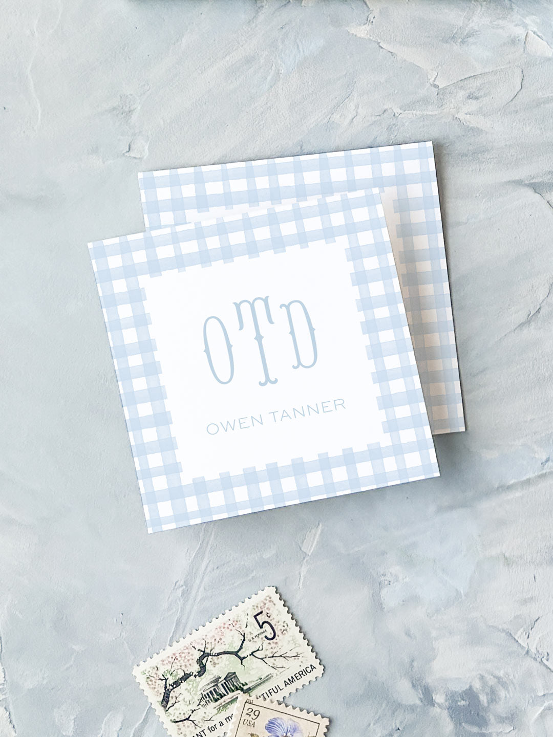 Two elegant blue gingham calling cards with monogram initials and a personalized name, displayed on a textured surface with vintage postage stamps nearby.