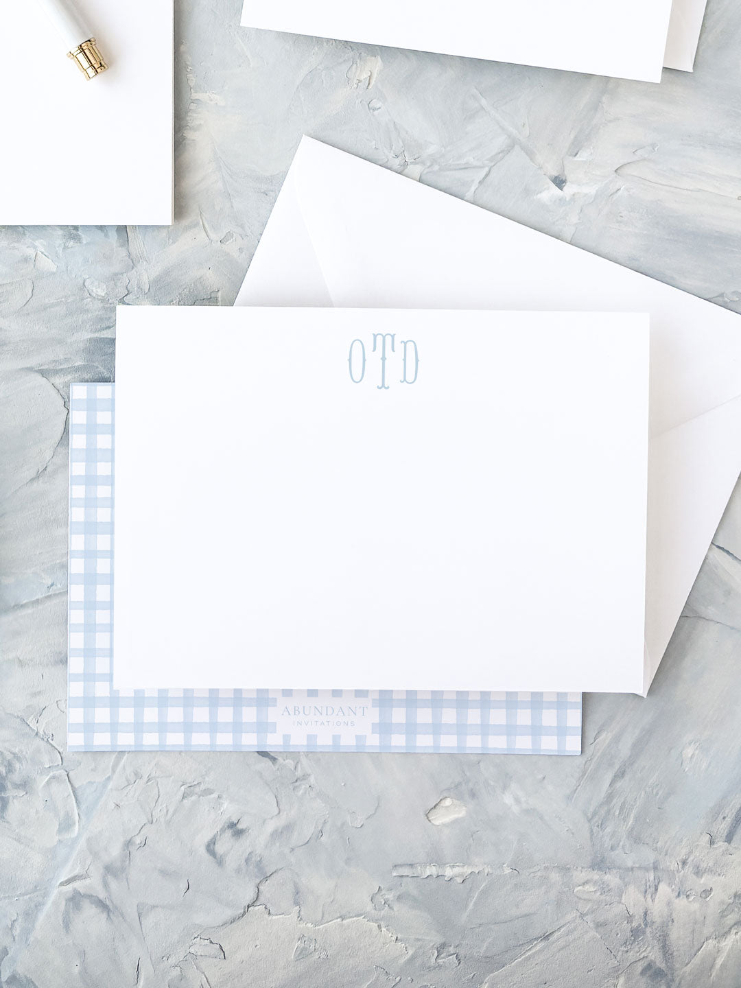 White flat notecard with light blue monogram lettering, paired with a matching blue gingham patterned envelope on a textured stone surface.
