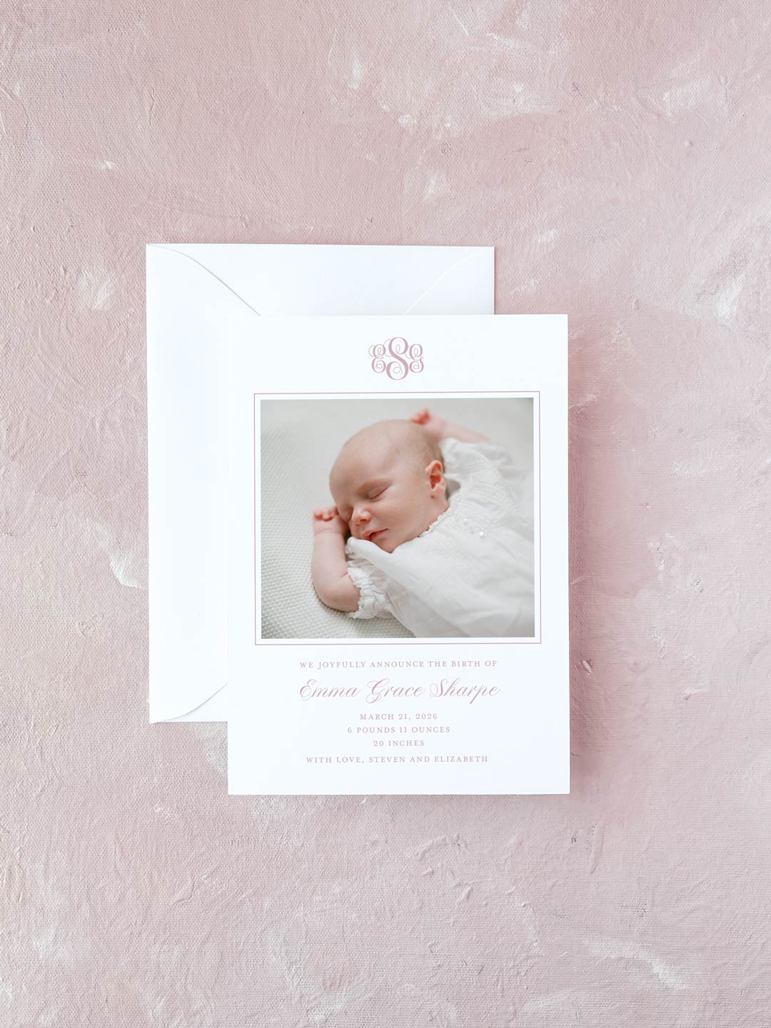 Elegant pink monogrammed birth announcement card featuring a photo of a sleeping newborn, presented with a matching white envelope on a soft pink background.