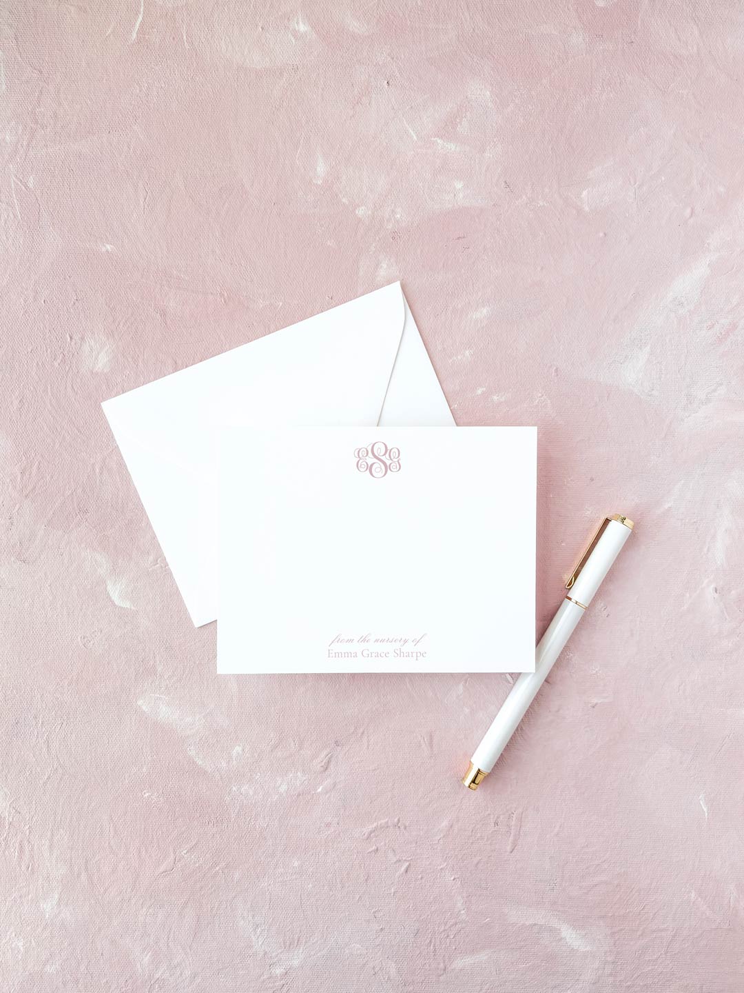 Elegant flat notecard with a delicate pink monogram on crisp white paper, paired with a matching envelope and gold-accented pen on a soft pink textured background.