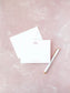 Elegant flat notecard with a delicate pink monogram on crisp white paper, paired with a matching envelope and gold-accented pen on a soft pink textured background.