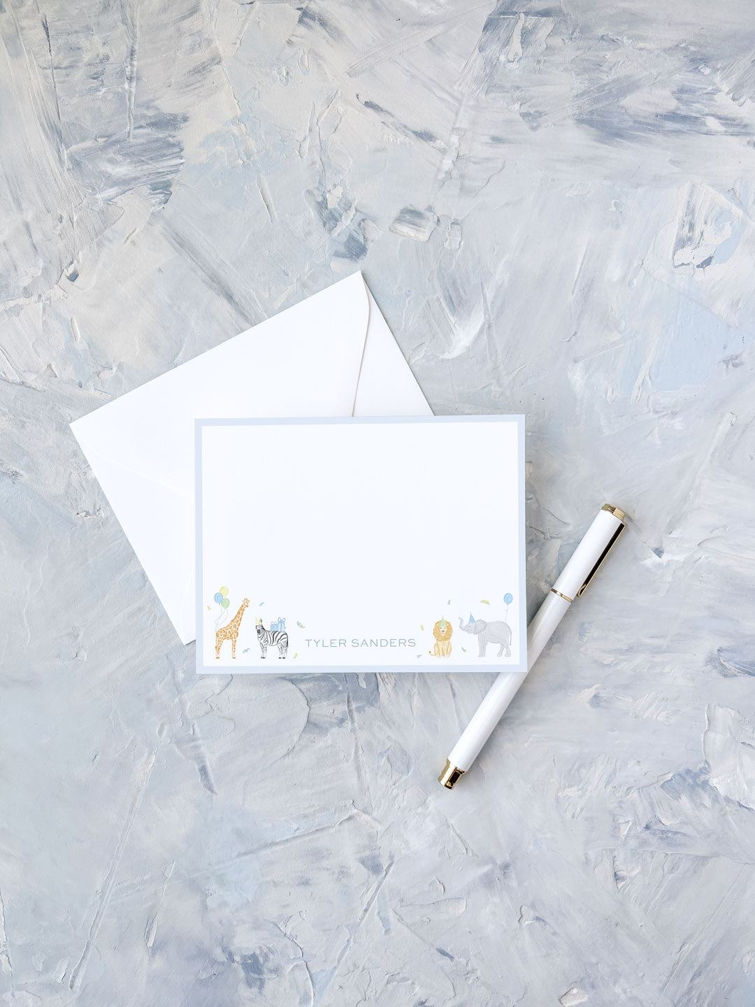Personalized flat notecard with playful animal illustrations and light blue border, accompanied by a white envelope and elegant pen on textured background.