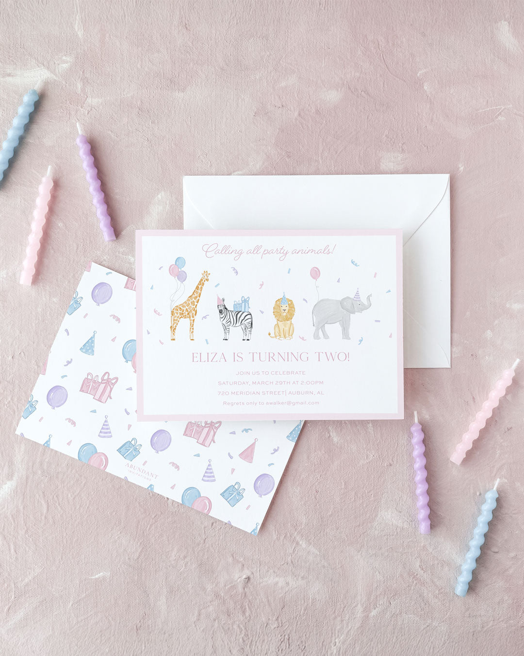 Pink birthday invitation with illustrated safari animals and festive party-themed envelope on a soft pink background.