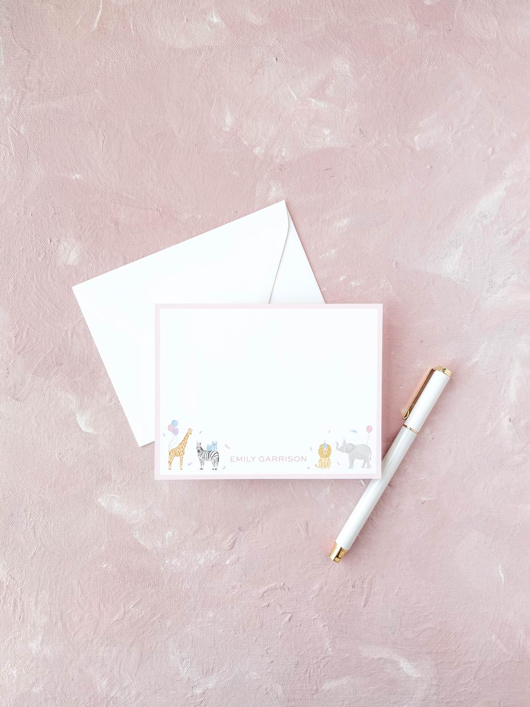 White flat notecard with pink border featuring cute watercolor animal illustrations and personalized name at the bottom, placed next to a white envelope and pen on a textured pink surface.