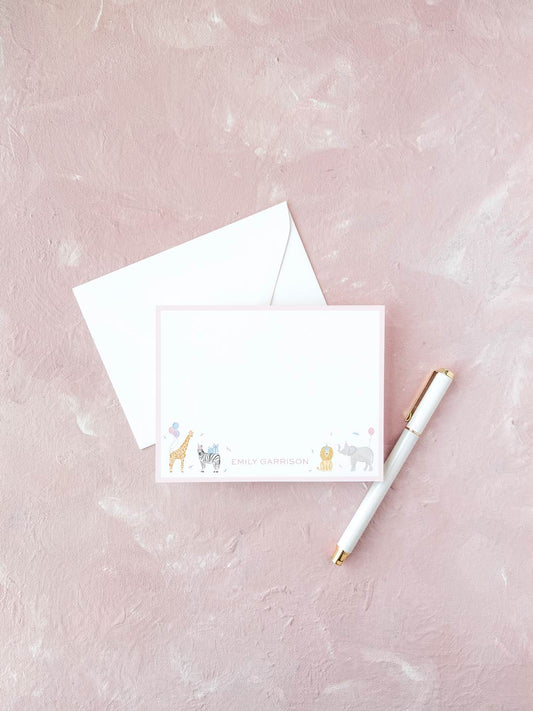 White flat notecard with pink border featuring cute watercolor animal illustrations and personalized name at the bottom, placed next to a white envelope and pen on a textured pink surface.