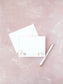 White flat notecard with pink border featuring cute watercolor animal illustrations and personalized name at the bottom, placed next to a white envelope and pen on a textured pink surface.