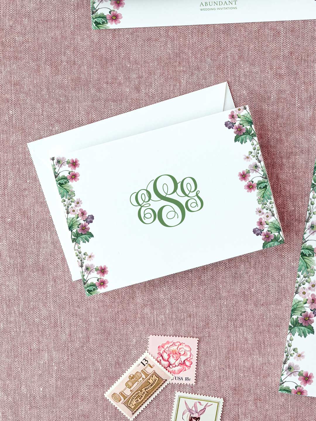 Elegant folded notecard with green monogram and floral border on textured pink background alongside vintage stamps