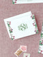 Elegant folded notecard with green monogram and floral border on textured pink background alongside vintage stamps