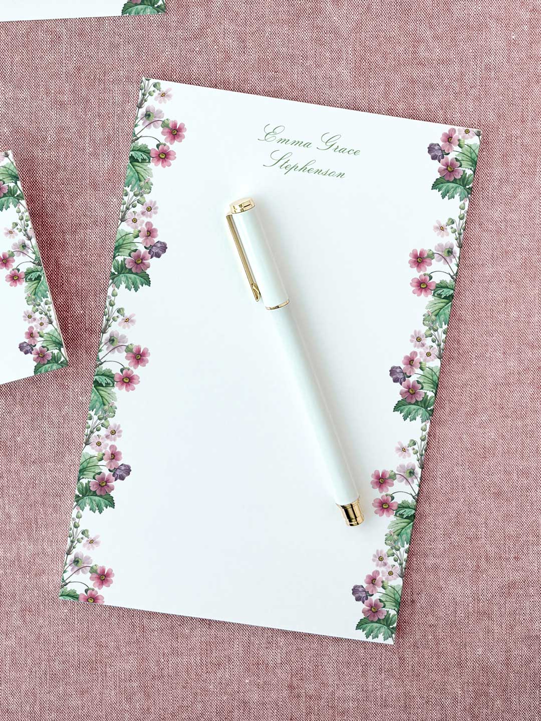 Notepad with floral border and elegant script, featuring a white pen with gold accents on a textured pink background