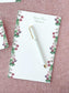 Notepad with floral border and elegant script, featuring a white pen with gold accents on a textured pink background
