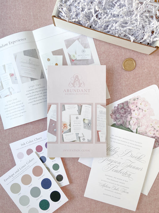 A neatly arranged sample pack showing personalized stationery pieces, including an invitation guide, ink color chart, and elegant wedding invitation designs on a soft pink background.