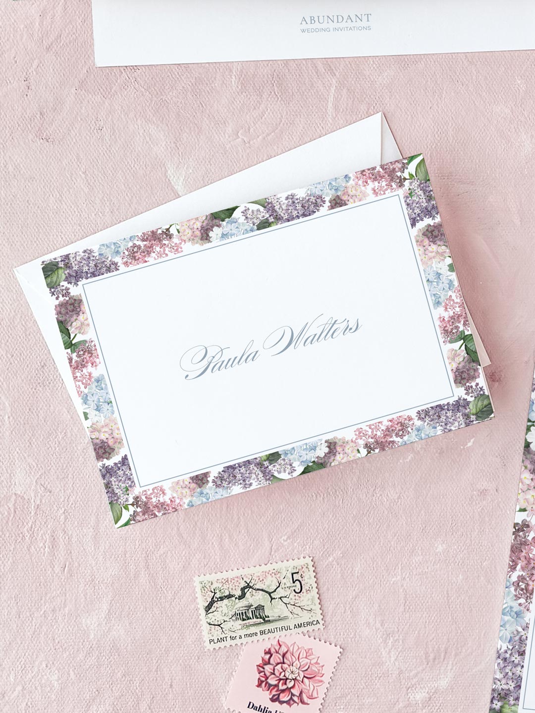 Folded notecard with delicate watercolor floral border on soft pink background, personalized elegant calligraphy name center.