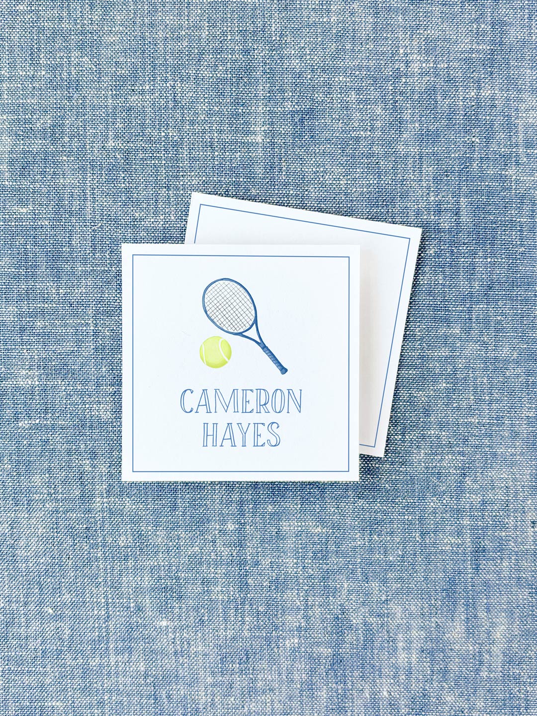 Square personalized calling card featuring a tennis racket and ball illustration with a classic serif name printed below on a blue textured fabric background.