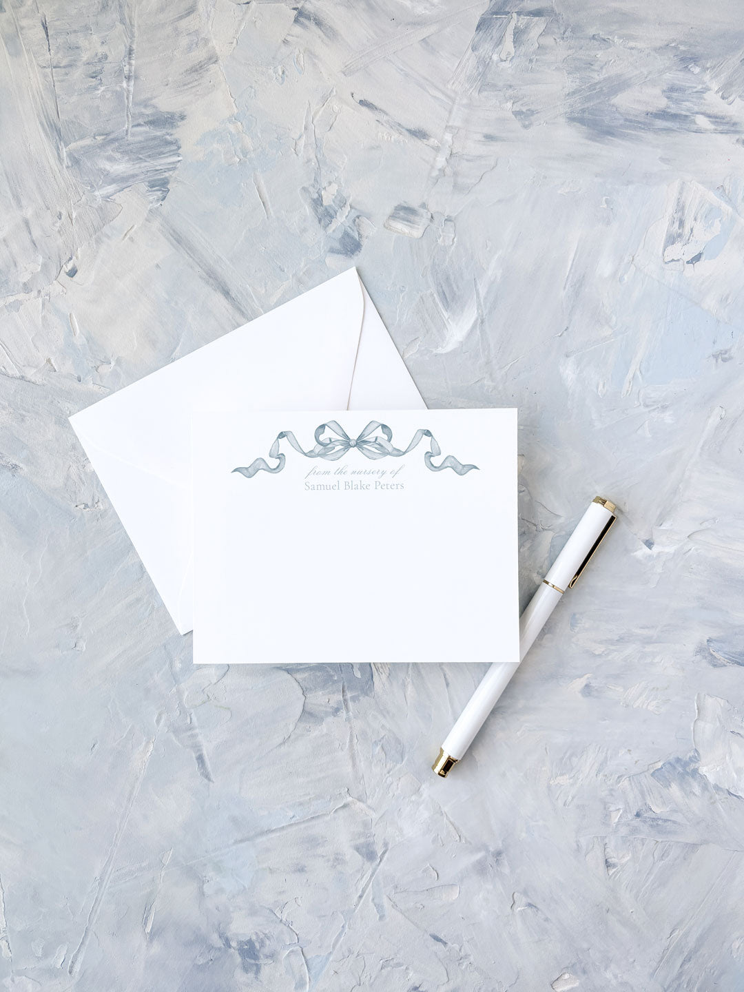 Elegant flat notecard featuring a delicate blue watercolor bow design, paired with a matching envelope and white pen on a textured surface.