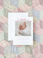 Birth announcement card featuring a sleeping baby dressed in white, decorated with a delicate pink watercolor bow on a soft pastel patchwork quilt background.