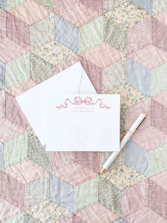 White flat notecard featuring a delicate pink watercolor bow design on top, placed on a pastel-colored quilt with an elegant pen nearby.